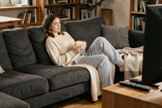 Jeune femme confortablement affalée dans un grand canapé sectionnel gris avec un bol de popcorn en regardant Netflix